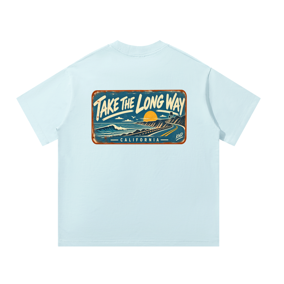 Take the Long Way Tee, Sky Blue — back print: a coast road winding along the cliffs at sunset past a breaking wave, in a weathered “Take the Long Way · California” sign