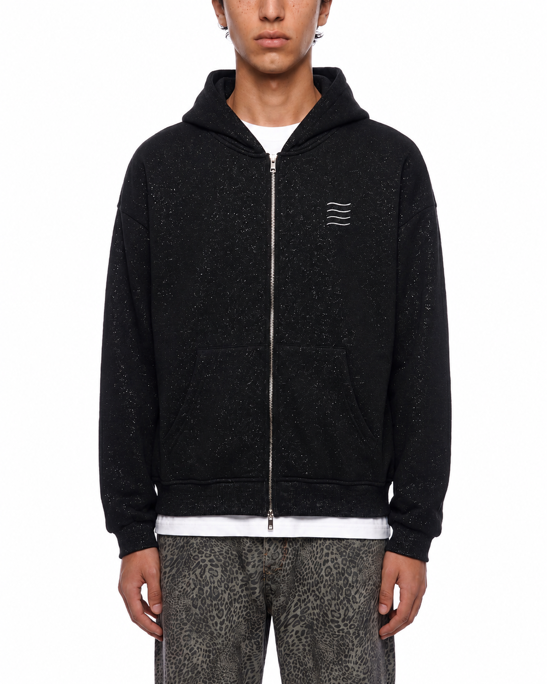 A model wearing a black zip-up hoodie with a subtle speckled texture, featuring a small white three-stripe logo on the left chest, styled over a white t-shirt and patterned gray pants.