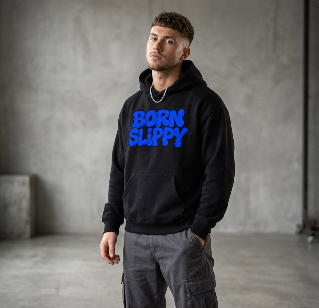 Born Slippy hoodie worn by a model against a concrete wall — black oversized pullover with BORN SLIPPY across the chest in blue bubble lettering, hand in the kangaroo pocket