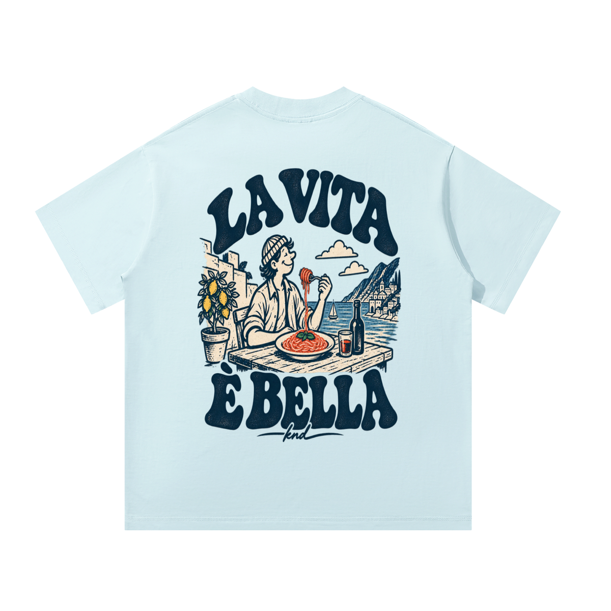 La Vita È Bella Tee, Sky Blue — back print: a man twirling spaghetti at a seaside table with a lemon tree and red wine, under “La Vita È Bella” lettering