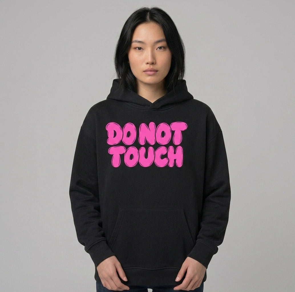 Do Not Touch hoodie worn by a model — black oversized pullover with DO NOT TOUCH across the chest in pink bubble lettering, hood down and kangaroo pocket visible