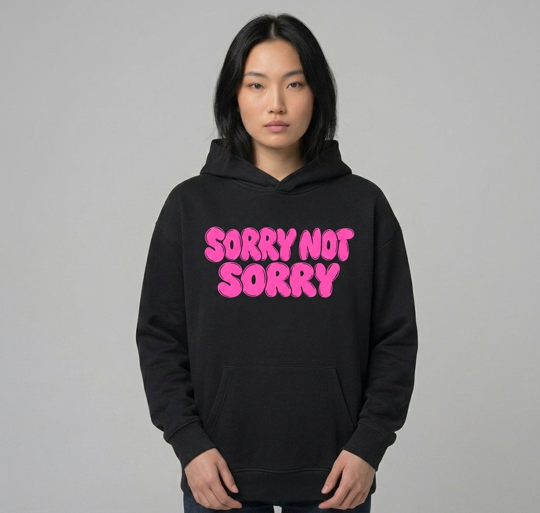 Sorry Not Sorry hoodie worn by a model — black oversized pullover with SORRY NOT SORRY across the chest in pink bubble lettering, hood down